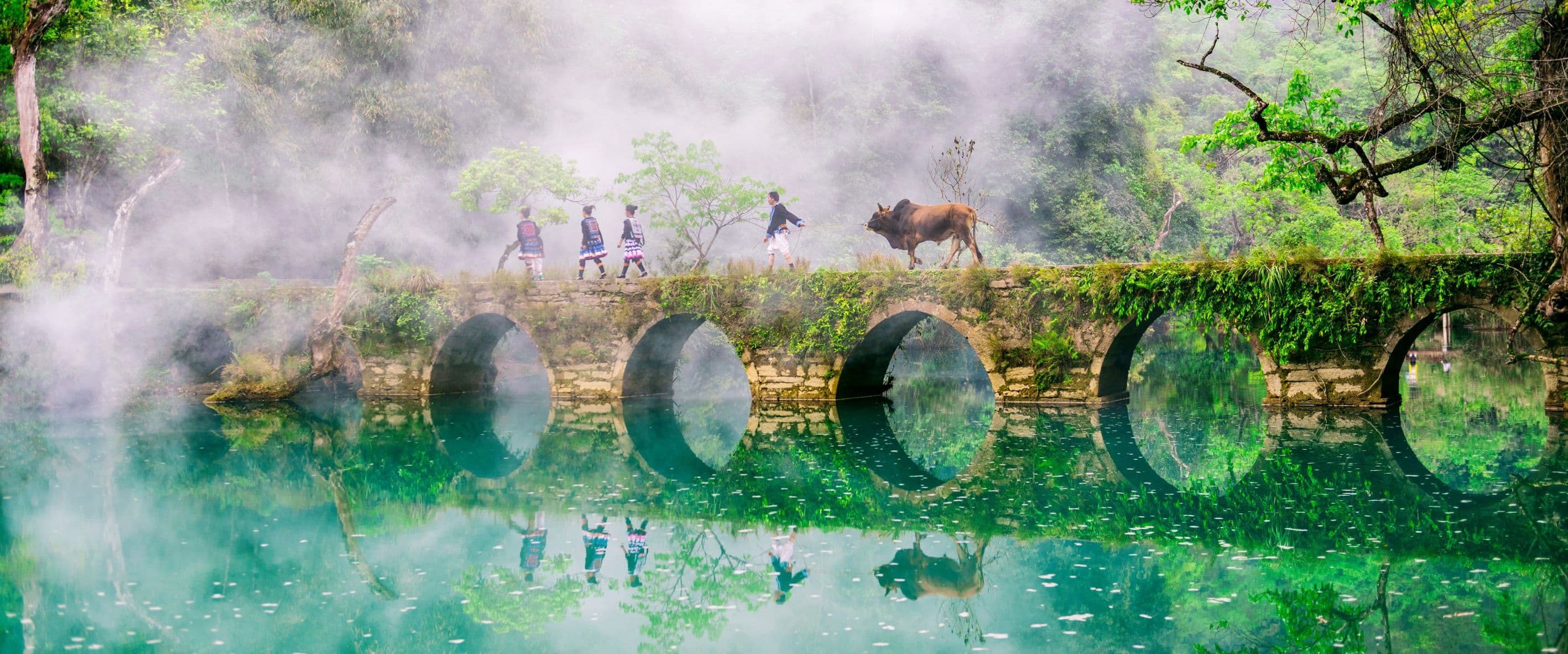 A stone bridge reflected in turquoise water beneath the mist